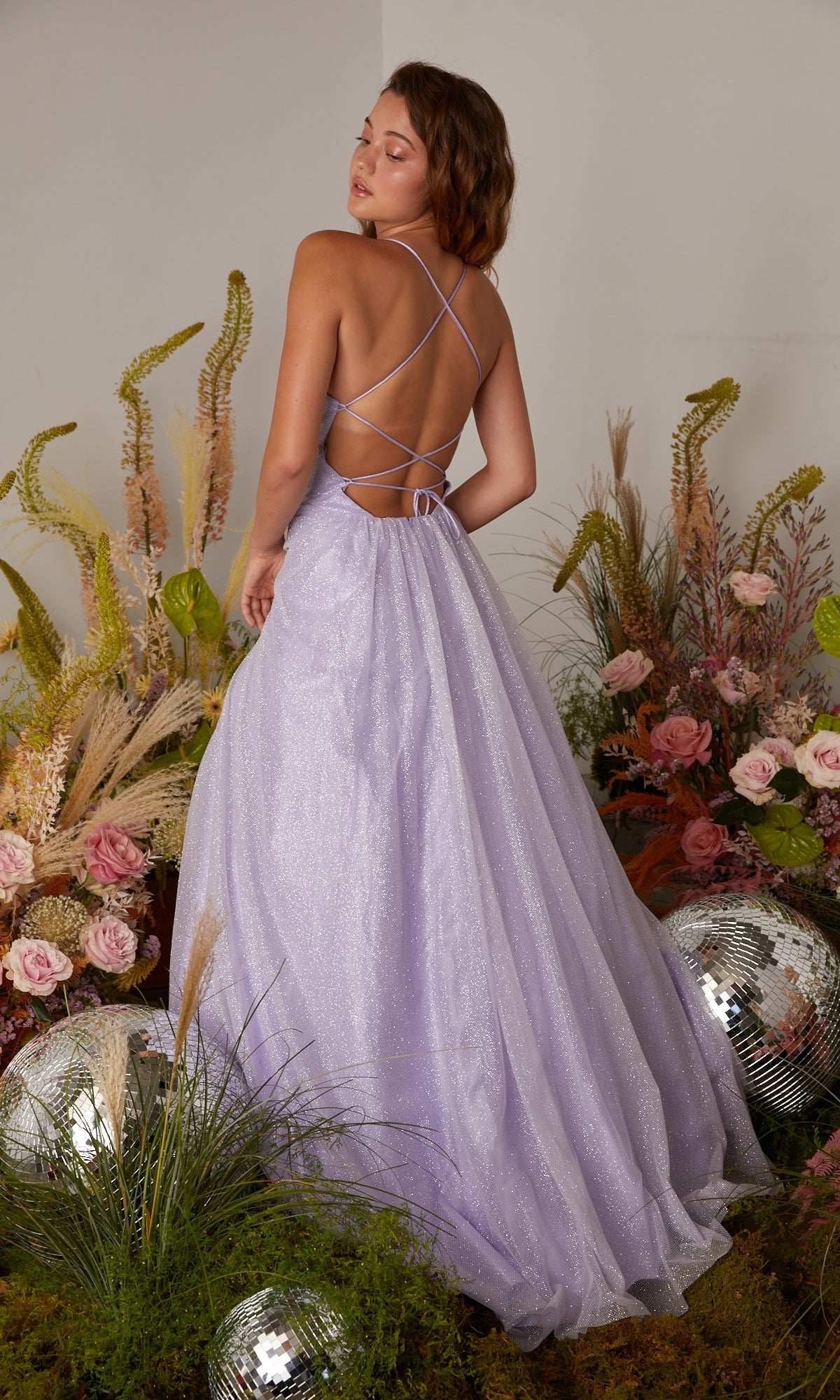 A woman wearing the Strappy-Back Glitter Prom Ball Gown: Eureka 9009 stands with her back to the camera, surrounded by flowers, greenery, and disco balls.